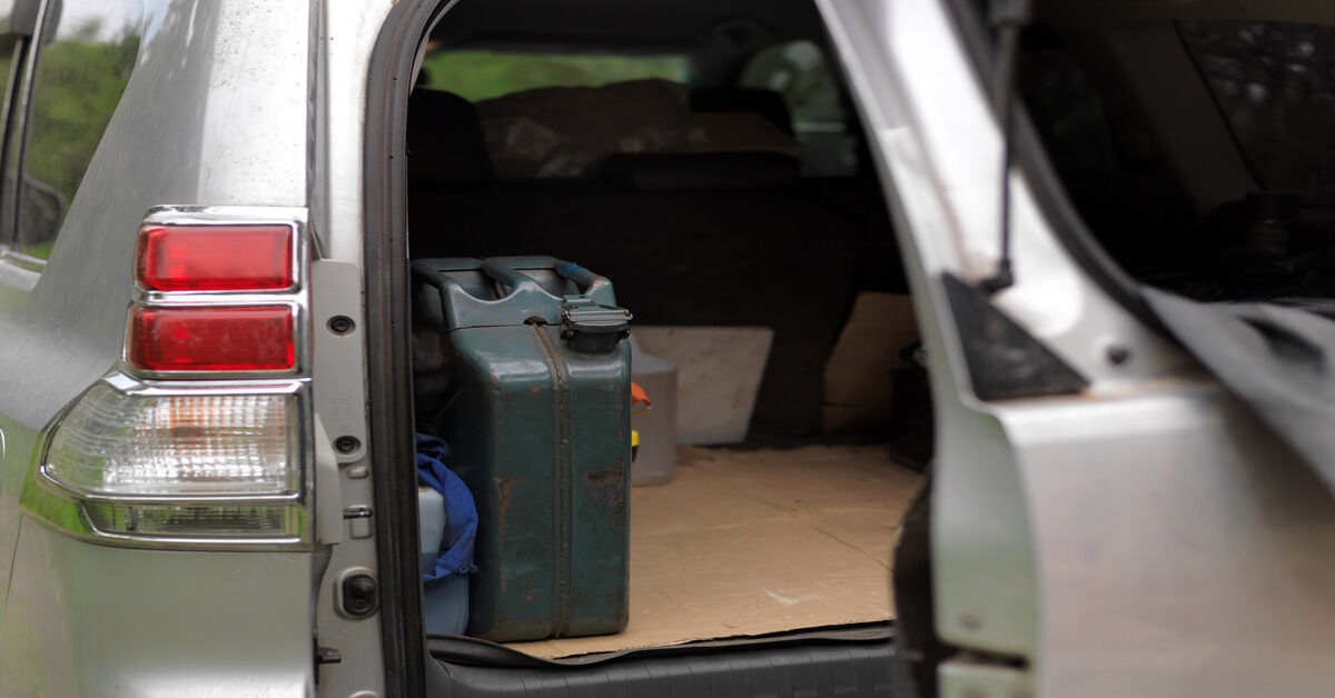 A dark green, classic metal jerry can in the back of a van's open trunk. The floor of the trunk is covered with cardboard.